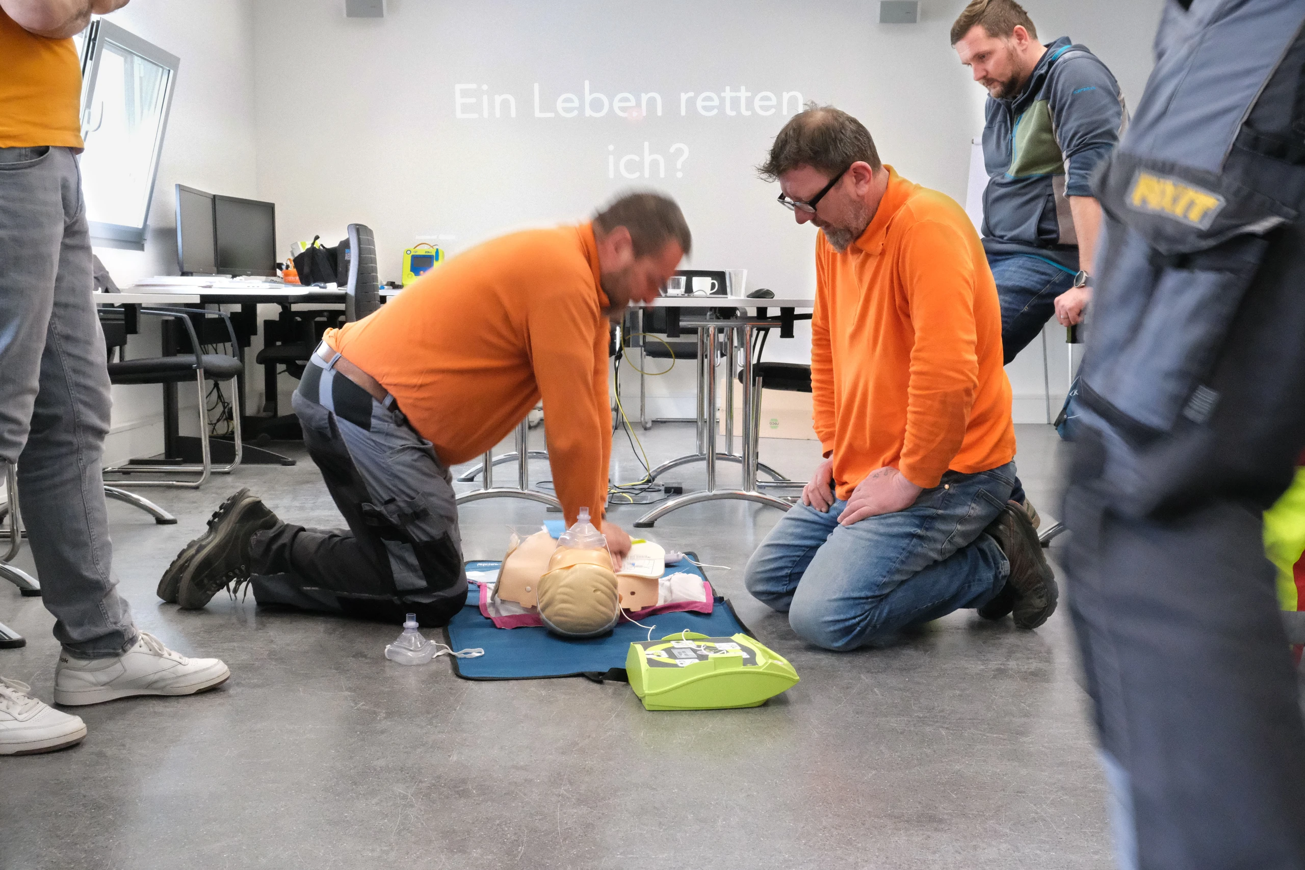 Deux employés de Fixit en t-shirt orange s'exercent à la réanimation sur un mannequin avec un défibrillateur.