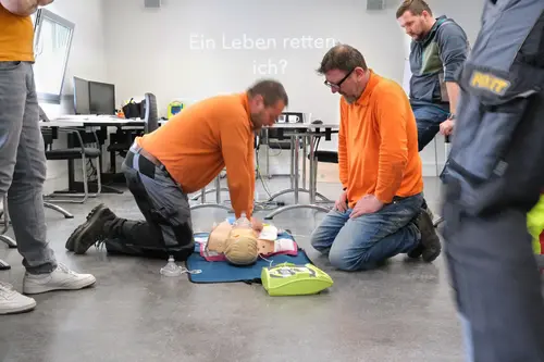 Deux employés de Fixit en t-shirt orange s'exercent à la réanimation sur un mannequin avec un défibrillateur.