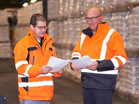 Zwei Fixit Mitarbeiter im Bereich Logistik in Warnkleidung stehen vor dem Produktlager mit Fixit Säcken und prüfen Lieferpapiere