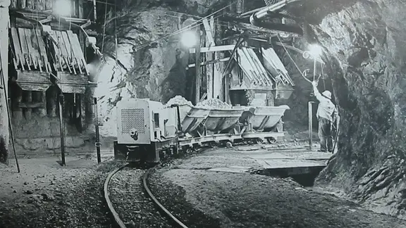 Schwarz-Weiss Fotografie aus einem Stollen mit Arbeiter, einem Zug aus Loren beladen mit Gesteinsbrocken und der Befüllungsanlage im Hintergrund