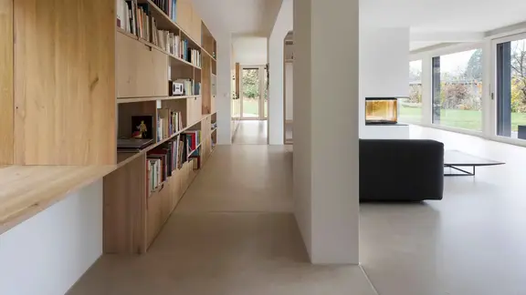 Moderner Wohnbereich mit grossem Holzregal für Bücher auf der linken Seite, einem schwarzen Sofa sowie Wänden und Decke mit Gipsputz