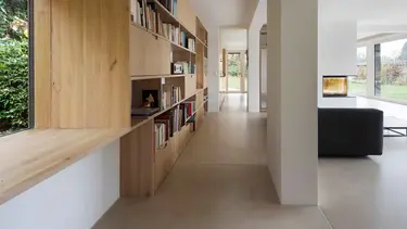 Salon moderne avec une grande étagère en bois pour les livres sur la gauche, une colonne en béton et un canapé noir, ainsi que des murs et un plafond en plâtre.